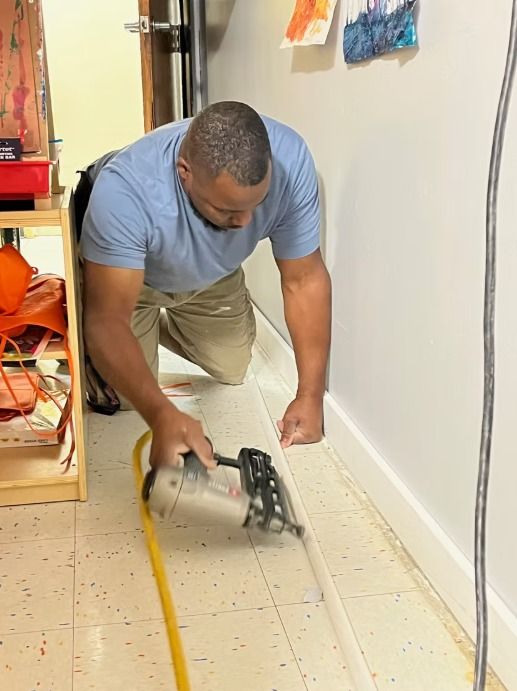 Man using nail gun on baseboard in a hallway. Yellow air hose visible.