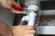 Hands using red pliers to fix a leaking white pipe under a sink