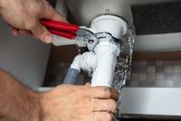 Hands using red pliers to fix a leaking white pipe under a sink
