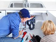 Two people inspect plumbing under a sink with tools in a white kitchen cabinet