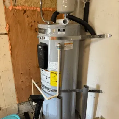 Gray water heater strapped to plywood wall in a utility closet, with pipes and warning labels.