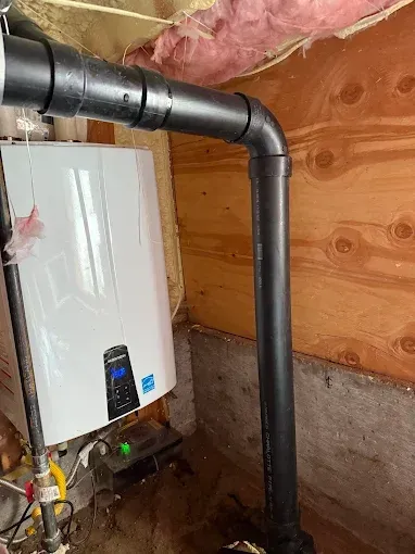 White boiler with black vent piping in a basement utility area, next to wood and concrete walls