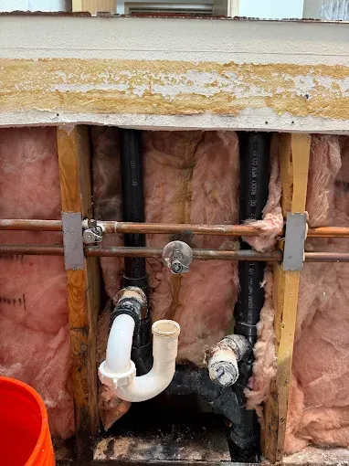 Exposed bathroom plumbing in a wall cavity with pink insulation and white PVC pipes under a sink drain.
