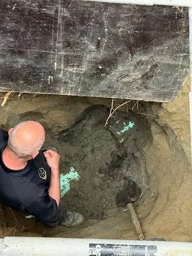 Person crouches in a dirt excavation under a concrete slab beside a green pipe.