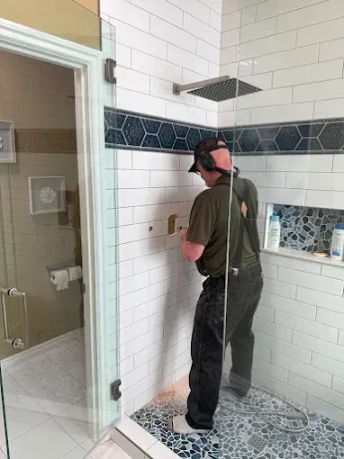 Man in a tiled shower stall cleaning white wall tiles with a spray bottle and cloth