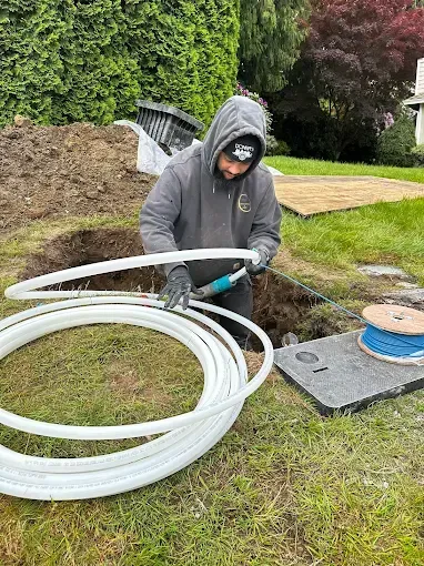 Worker in gray hoodie installing white coiled tubing in a grassy yard beside a trench.