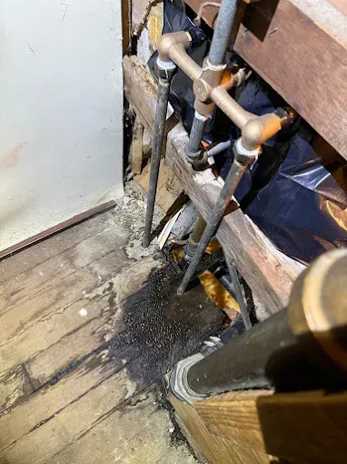 Damaged floor beside exposed pipes and debris near a doorway
