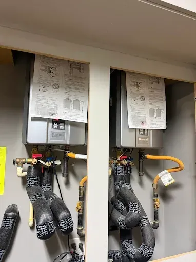 Two wall-mounted utility meters with hoses and labels in a service closet, one with orange tubing.