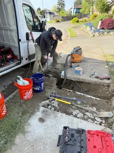 Worker repairing a sidewalk trench beside a van, with tools and buckets on a residential street