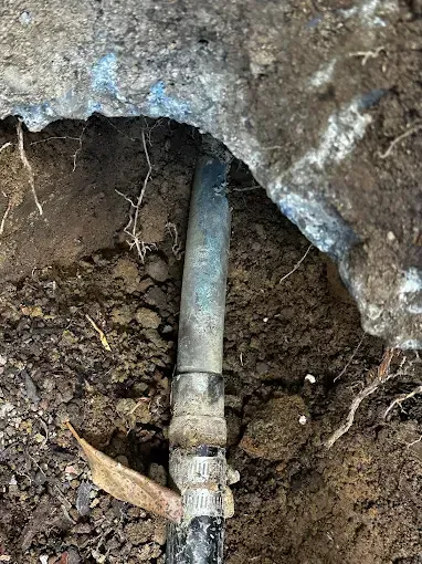 Exposed blue-gray pipe with a clamp under a concrete slab in a dirt excavation