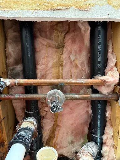 Exposed plumbing in a wall cavity with pink insulation, black pipes, and copper pipes with fittings