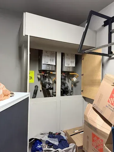 Cluttered utility room with a white cabinet, exposed pipes behind glass doors, and stacked cardboard boxes.