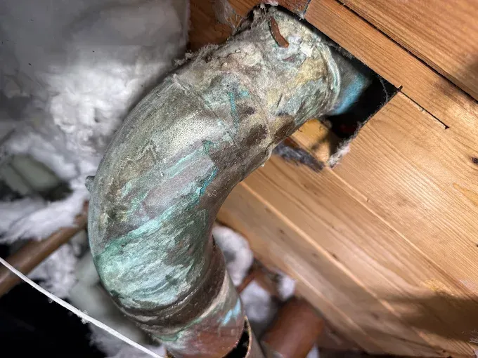 Corroded metal pipe elbow entering a wooden wall near white insulation