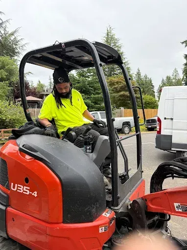 Worker in neon yellow jacket operating a red mini excavator outdoors.