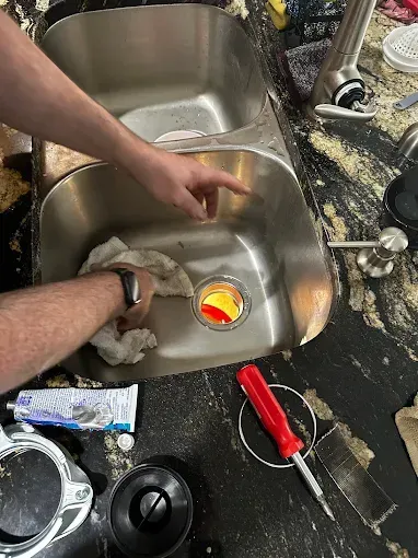 Person cleaning a cluttered kitchen sink with hands and sponge, red-handled knife nearby