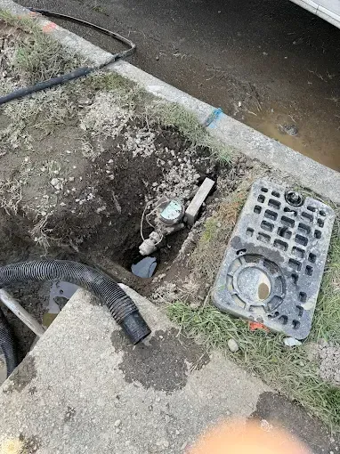 Open utility hole beside a driveway, with exposed pipes and a black grate nearby.