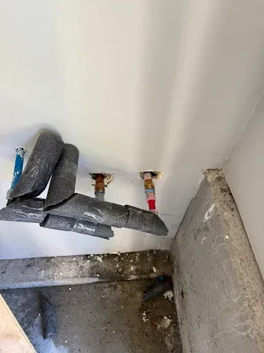 Exposed ceiling pipes with black insulation and a concrete wall in a damaged corner.