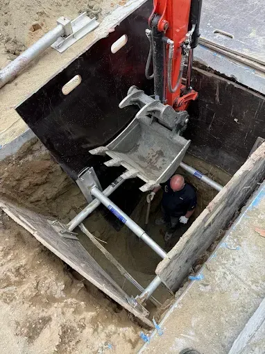 Worker inside a deep excavation beside a red excavator bucket and shoring boards
