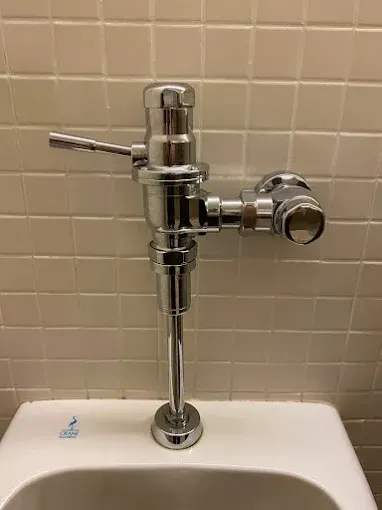 Chrome flushometer on a white toilet against beige tiled wall