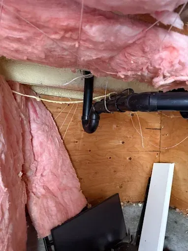 Pink insulation around black plumbing pipes beneath a floor joist, with exposed wood and debris.