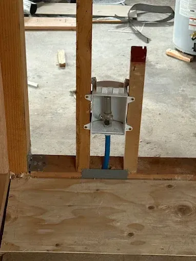 Construction framing with a mounted metal plumbing fixture between wooden studs on a plywood floor