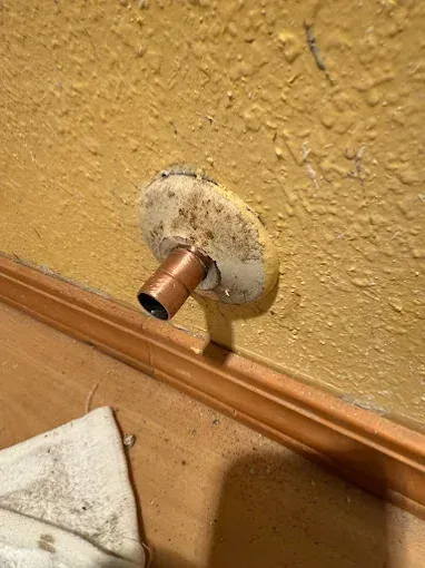 Copper pipe stub protruding from a beige textured wall above a wood baseboard.