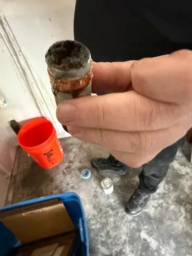 Hand holding a rusty pipe fitting over a dusty floor with an orange bucket nearby