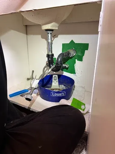 Blue and silver plumbing fixture inside a cabinet, with pipes and a green tape patch on the wall.