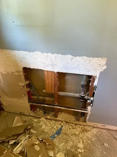 Damaged wall with a rough rectangular hole exposing pipes and wooden framing, debris on the floor