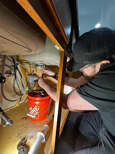 Worker crouches under a sink, using tools to repair plumbing beside an orange bucket.