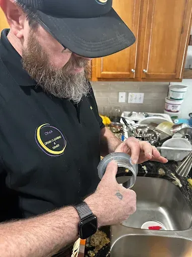 Man cleaning a cup at the kitchen sink, wearing a black cap and shirt.
