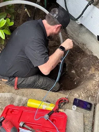 Technician in black clothing inspects a ground hole with a blue cable beside red equipment outdoors