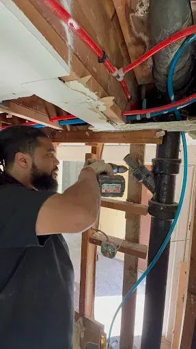 Man drilling into ceiling joist beside exposed pipes and wires in unfinished room