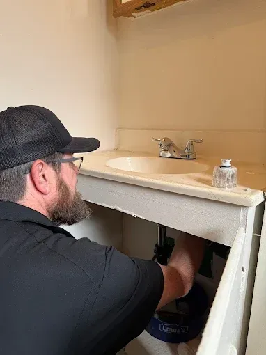 Person repairing plumbing under a bathroom sink, with tools and exposed pipes visible.
