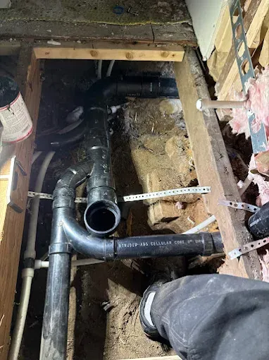 Plumbing repair under a floor: black pipes, dirt, and a measuring tape in an exposed crawlspace