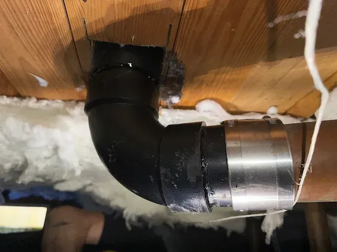 Black insulated HVAC duct elbow with metal clamp, mounted through a wooden ceiling with white insulation nearby