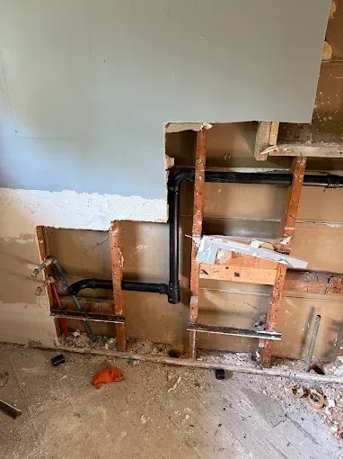 Wall with exposed plumbing and studs, showing drywall removal and a damaged lower section during renovation.