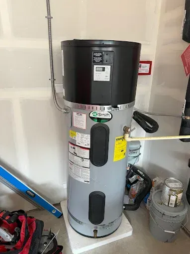 Tall gray water heater tank in a garage, with labels and pipes beside tools and supplies.