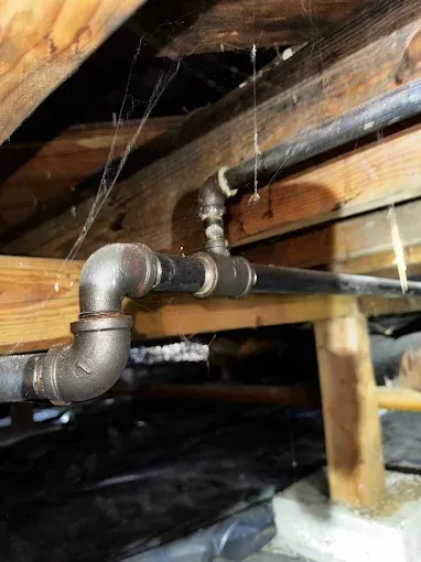 Old galvanized pipe elbow and black conduit under exposed wooden floor joists, with spider webs.