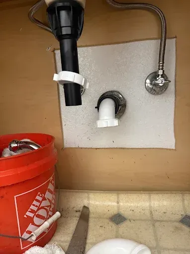 Under-sink plumbing with exposed pipes, a red Home Depot bucket, and a wrench on a tiled floor.
