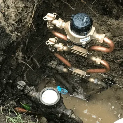 Exposed water meter and copper pipes in a muddy excavation hole