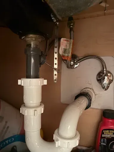 Under-sink plumbing with white PVC drain pipes, a chrome supply line, and a red cleaner bottle in the cabinet.