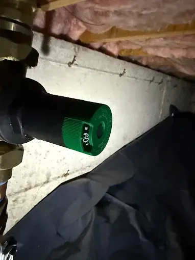 Green-capped pipe valve in a crawlspace next to insulation and black sheeting.