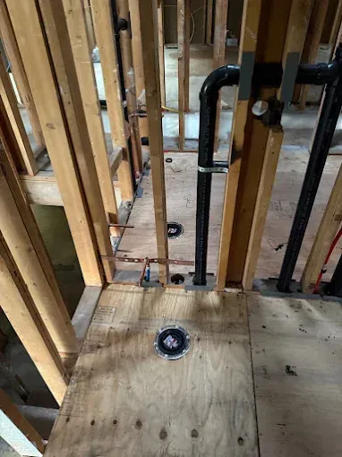 Unfinished bathroom framing with exposed pipes and a toilet flange on plywood subfloor.