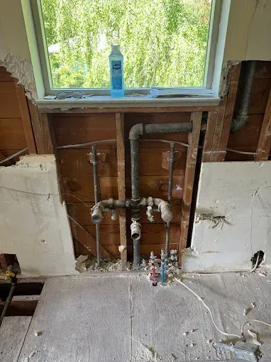 Bathroom wall under renovation with exposed plumbing beneath a window, showing pipes and unfinished drywall.