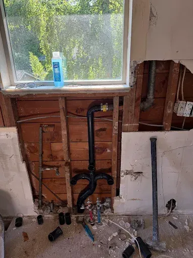 Partially demolished bathroom wall under a window, exposing plumbing and framing during renovation