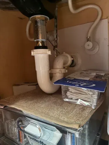 Under-sink plumbing with white drain pipes above a cabinet shelf holding a blue-labeled package.