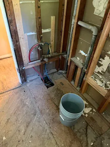 Exposed plumbing in a stripped wall with a gray bucket on a bare wood floor.
