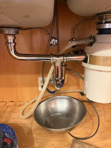 Under-sink plumbing with a metal drain pipe, beige disposal unit, and a bowl on the cabinet floor.
