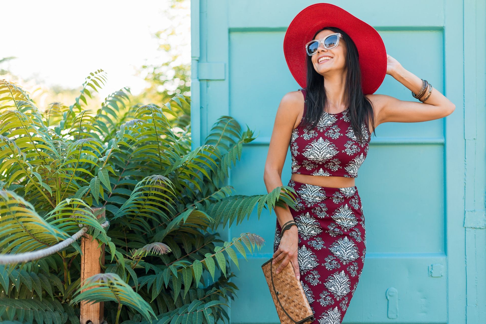 Mujer con sombrero rojo y traje estampado sonriendo cerca de una puerta verde azulado y vegetación.